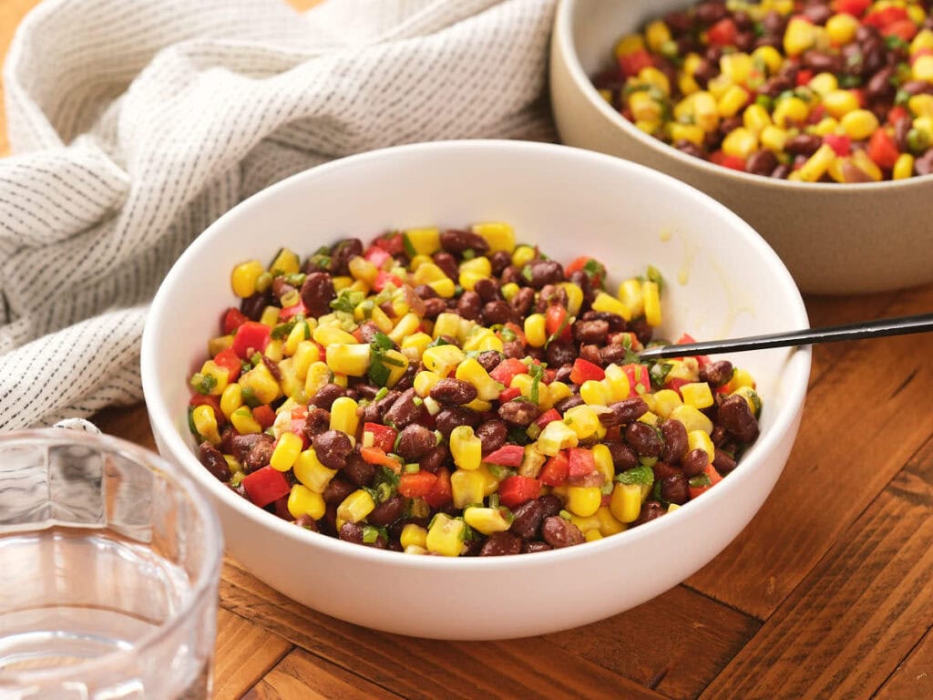 A bowl filled with a corn and black bean salad sits on a wooden table next to a glass of water and a striped cloth.