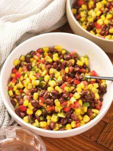 A white bowl filled with a colorful corn and black bean salad with a spoon inside and a striped cloth are nearby.