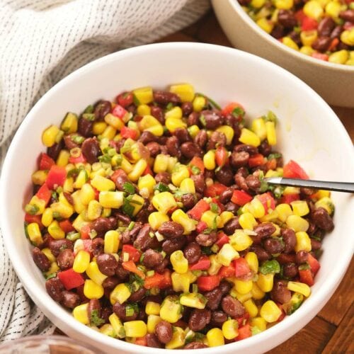 A white bowl filled with a colorful corn and black bean salad with a spoon inside and a striped cloth are nearby.