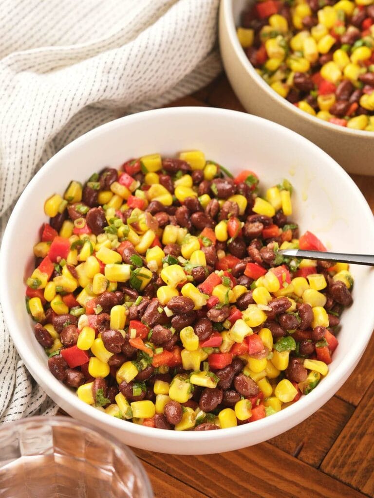 A white bowl filled with a colorful corn and black bean salad with a spoon inside and a striped cloth are nearby.