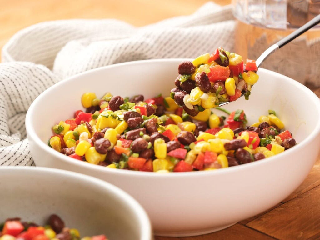 A bowl of corn and black bean salad and a spoon is scooping some salad from the bowl on a wooden surface.