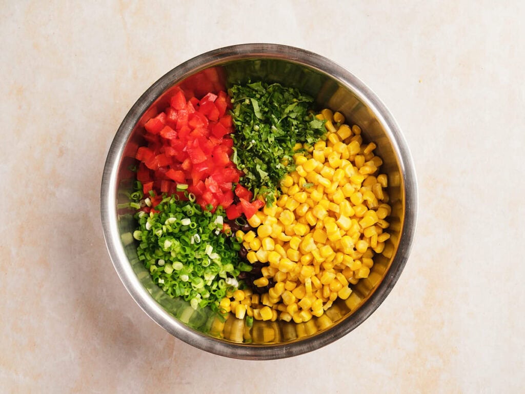 A metal bowl containing chopped bell peppers, cilantro, corn, and green onions arranged in separate sections on a light-colored surface.