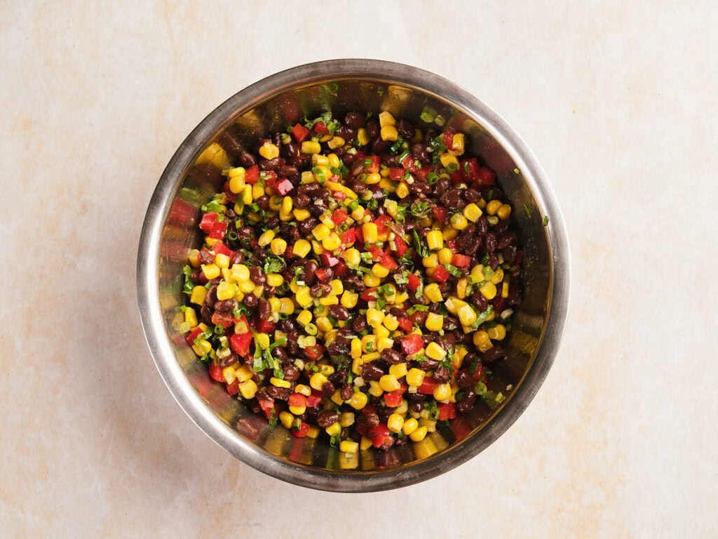A metal bowl filled with a colorful mixture of corn and black bean salad on a light surface.