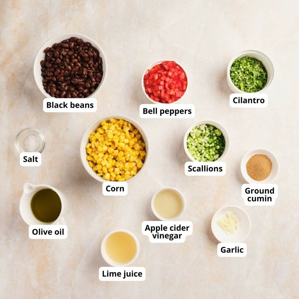 Top-down view of labeled ingredients in bowls of corn and black bean salad in a light surface.