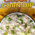 A bowl of creamy corn dip topped with chopped green onions, surrounded by yellow tortilla chips.