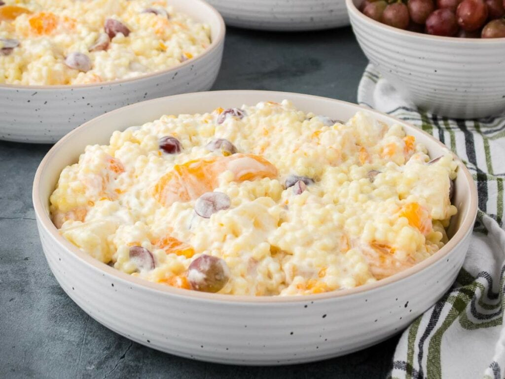 A white bowl filled with creamy frog eye salad on a table next to a striped cloth and a bowl of grapes.