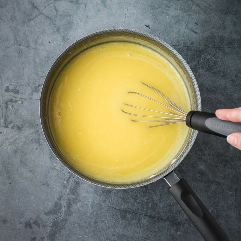 A hand is whisking a yellow liquid mixture in a saucepan on a dark grey countertop.