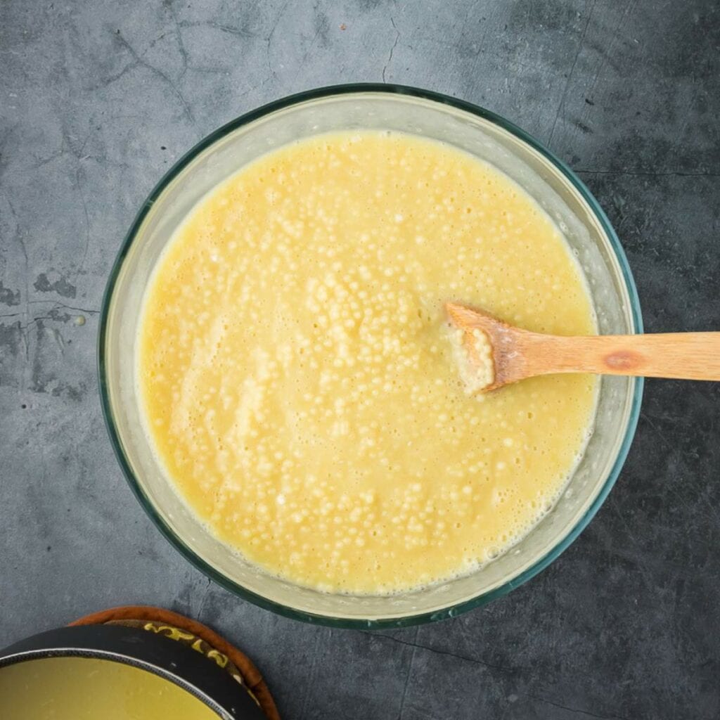A glass bowl pasta and custard sits on a gray countertop with a wooden spoon resting inside.