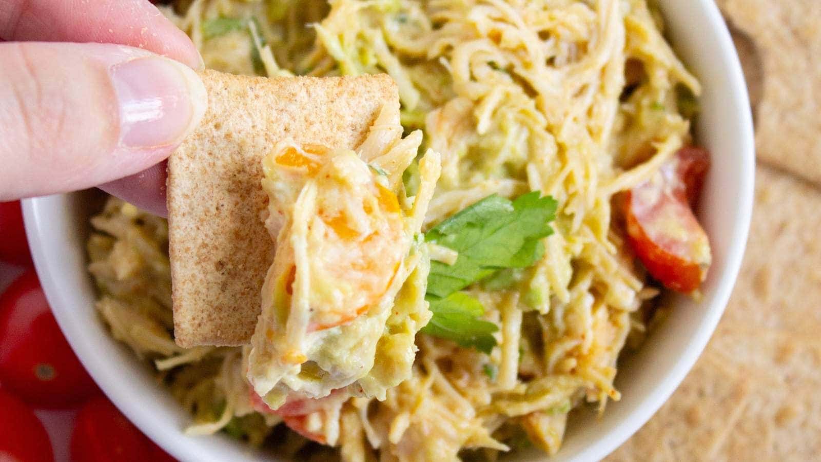 A hand holding a cracker topped with a healthy chicken salad, vegetables, and herbs.