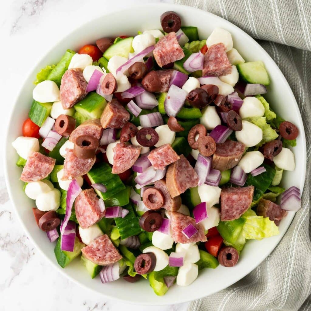 A bowl of salad with chopped lettuce, cucumbers, cherry tomatoes, red onions, salami, black olives, and mozzarella balls.
