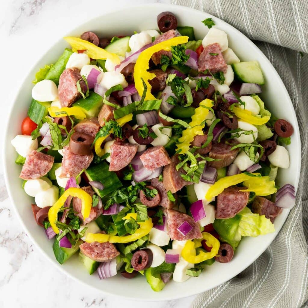 A bowl of chopped salad with lettuce, salami, mozzarella, olives, red onion, pepperoncini, cucumber, and fresh herbs.