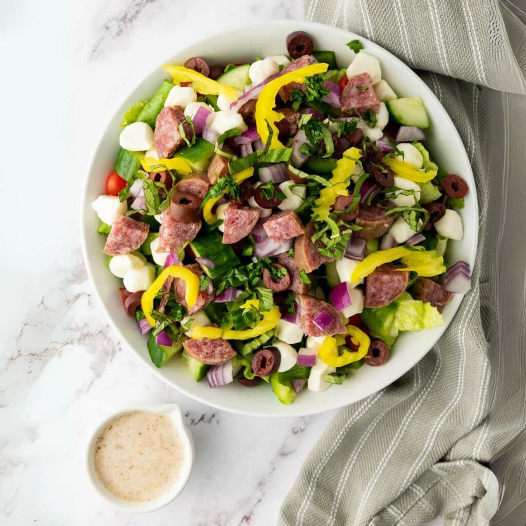 A bowl of salad with chopped lettuce, salami, mozzarella balls, red onions, olives, and pepperoncini.