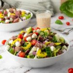 A bowl of Italian salad with cherry tomatoes, cucumbers, red onions, mozzarella balls, pepperoncini, and sliced salami, paired with a jar of dressing.