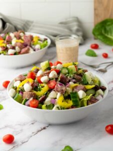 A bowl of Italian salad with cherry tomatoes, cucumbers, red onions, mozzarella balls, pepperoncini, and sliced salami, paired with a jar of dressing.