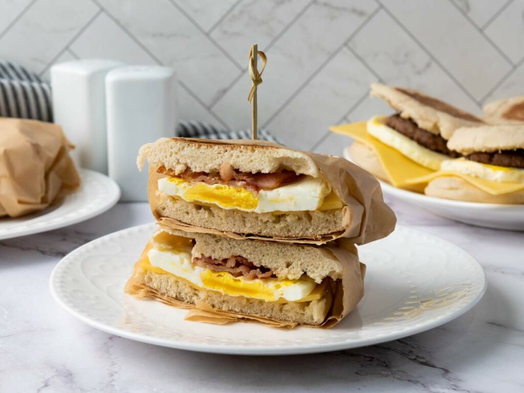 A make ahead breakfast sandwiches with egg, cheese, and meat is cut in half and stacked on a white plate, with more sandwiches in the background.