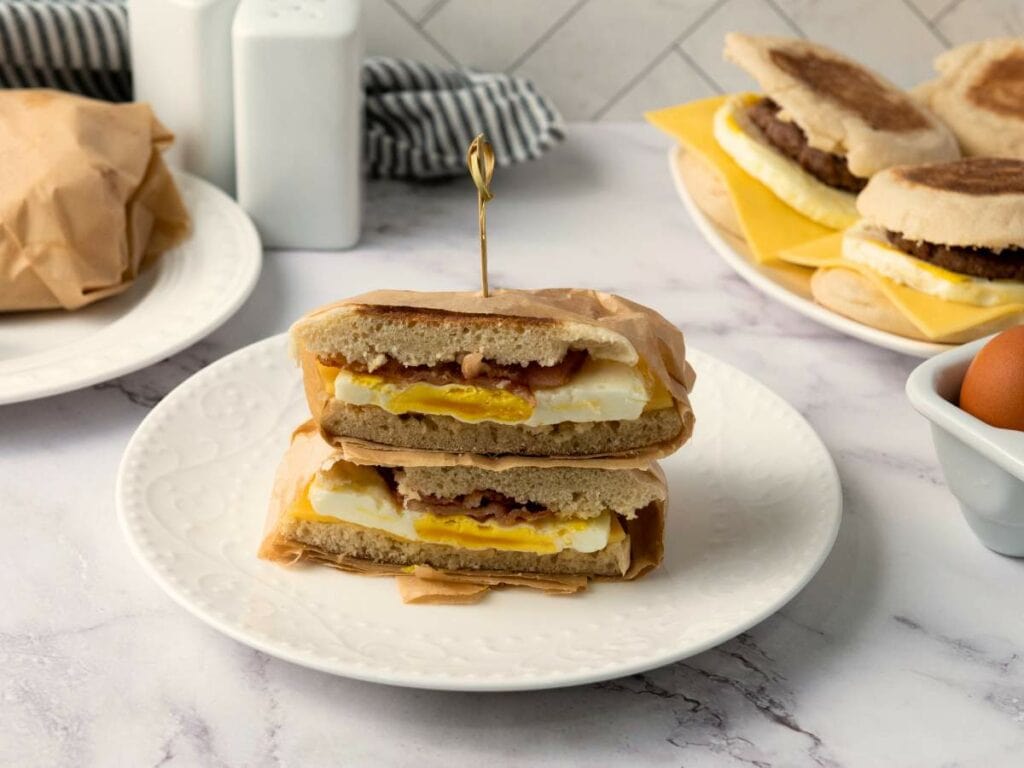 A make ahead breakfast sandwiches with egg, cheese, and bacon cut in half, stacked on a white plate.