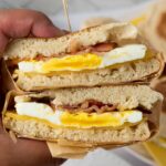 A hand holds a breakfast sandwich cut in half, showing layers of egg, bacon, and cheese.