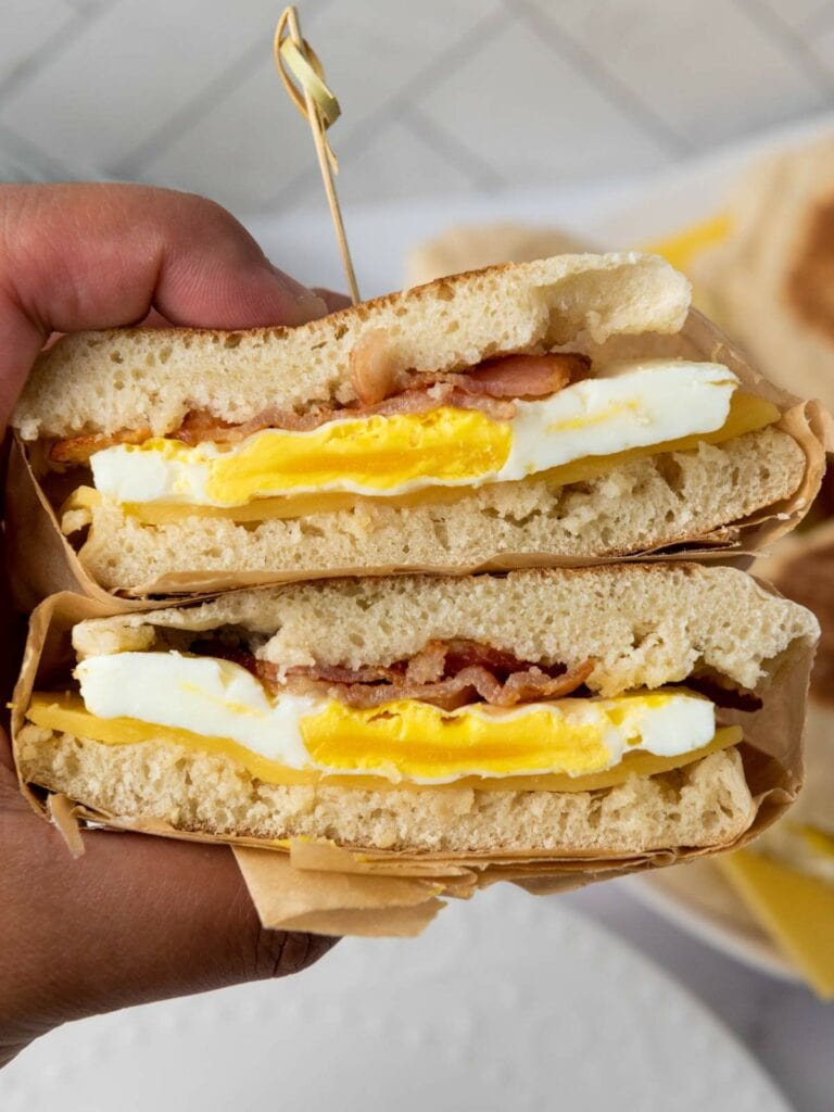 A hand holds a make ahead breakfast sandwich cut in half, showing layers of egg, bacon, and cheese.