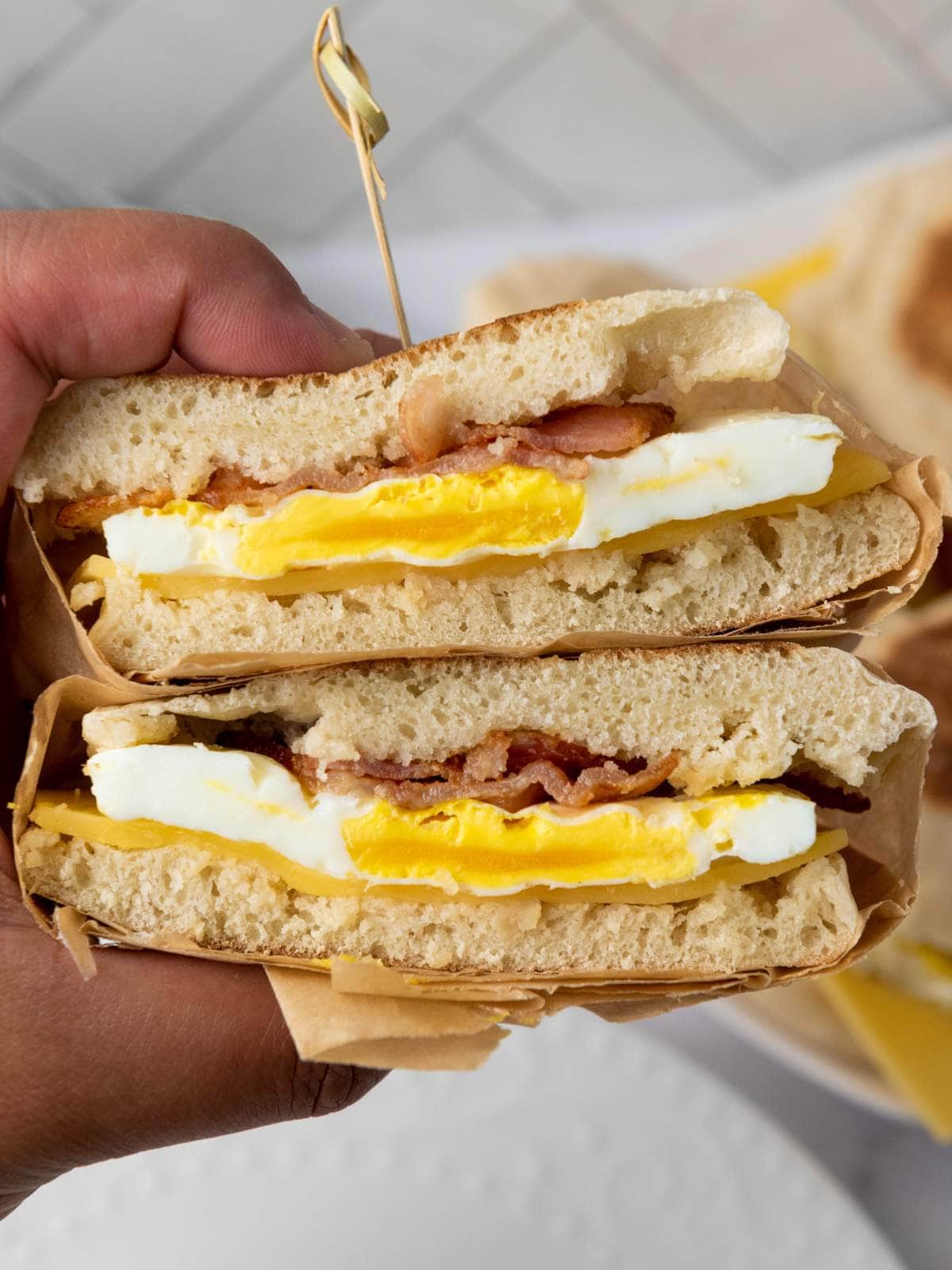 A hand holds a breakfast sandwich cut in half, showing layers of egg, bacon, and cheese.