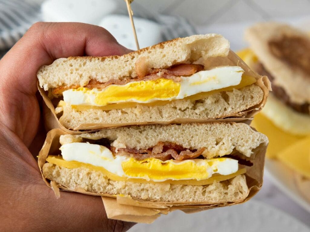 Hand holding a stacked make ahead breakfast sandwiches with bacon, egg, and cheese between two halves of ciabatta bread.