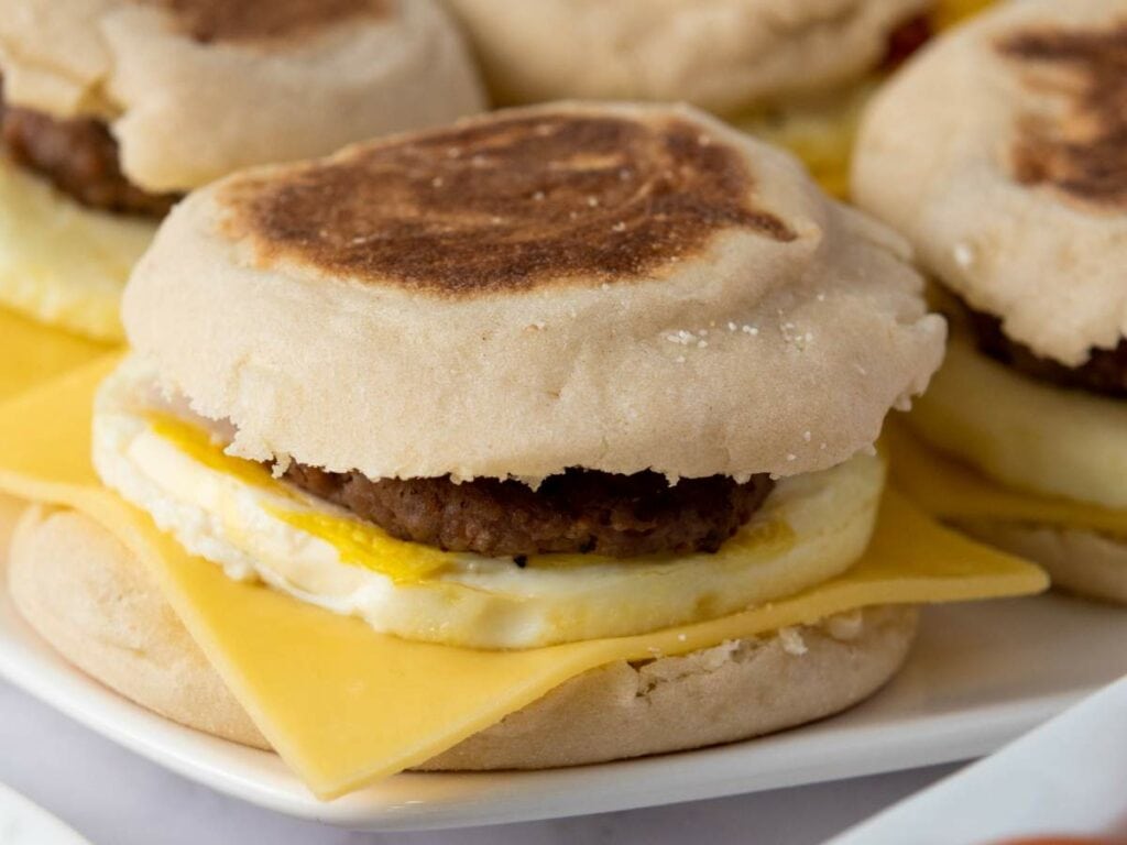 A make ahead breakfast sandwiches with egg, a slice of cheddar cheese, and a sausage patty on a white plate.