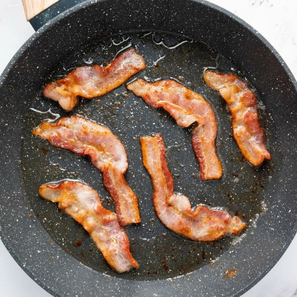 Six strips of cooked bacon in a black frying pan with visible grease.