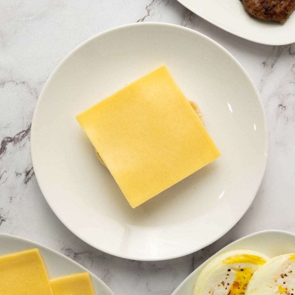 A slice of cheese on a plain muffin sits centered on a white plate, with other food items partially visible around the edges.