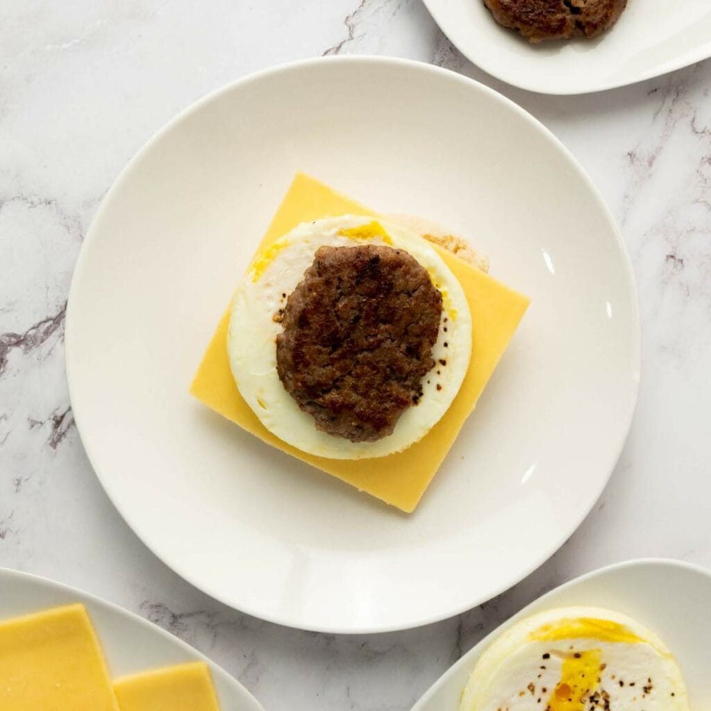 A muffin with a slice of cheddar cheese, a cooked egg, and a sausage patty on a white plate, viewed from above.