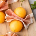 Skewers with slices of prosciutto e melone arranged on a wooden board, with sprigs of rosemary nearby.