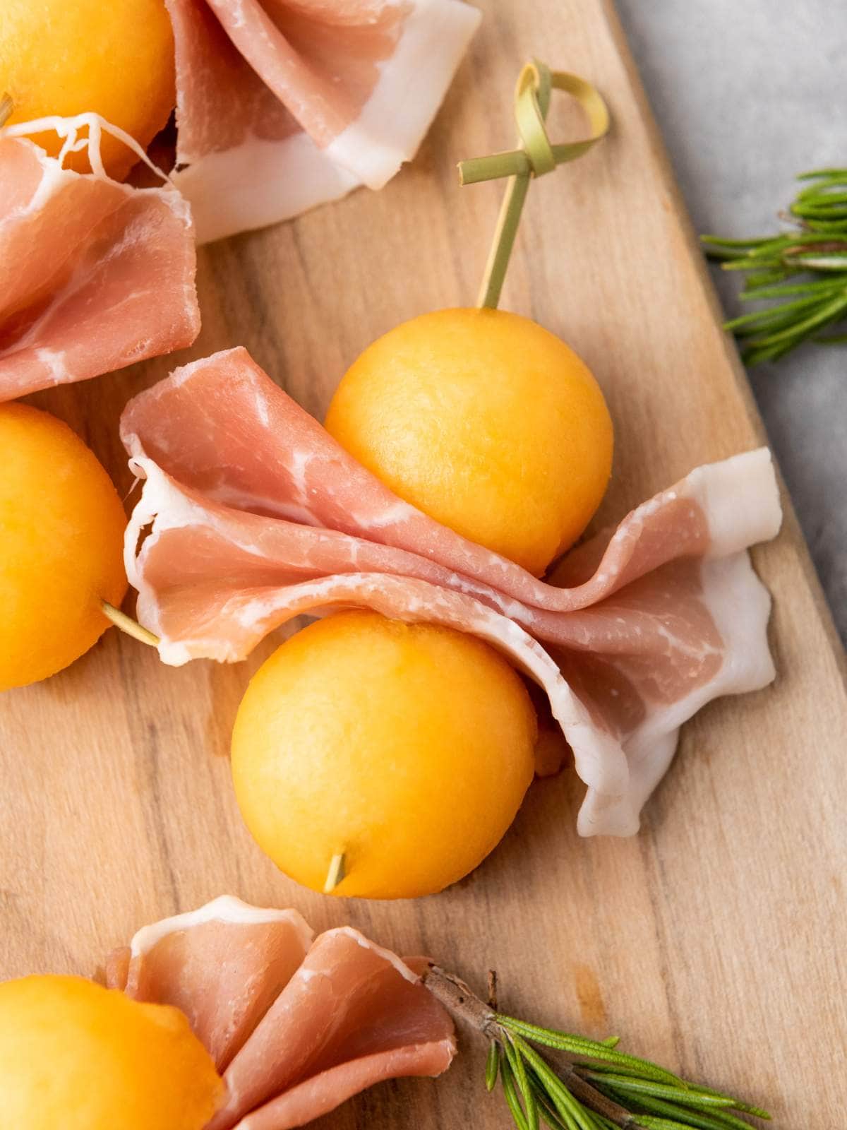 Skewers with slices of prosciutto e melone arranged on a wooden board, with sprigs of rosemary nearby.