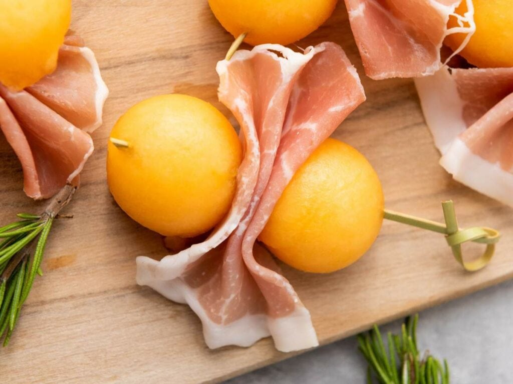 Skewered melon ball with prosciutto arranged on a wooden board with rosemary sprigs nearby.