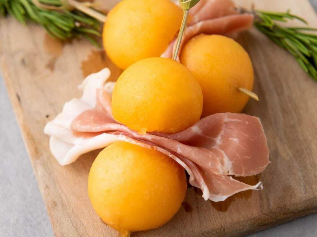 Four prosciutto e melone arranged on a wooden board, garnished with rosemary.