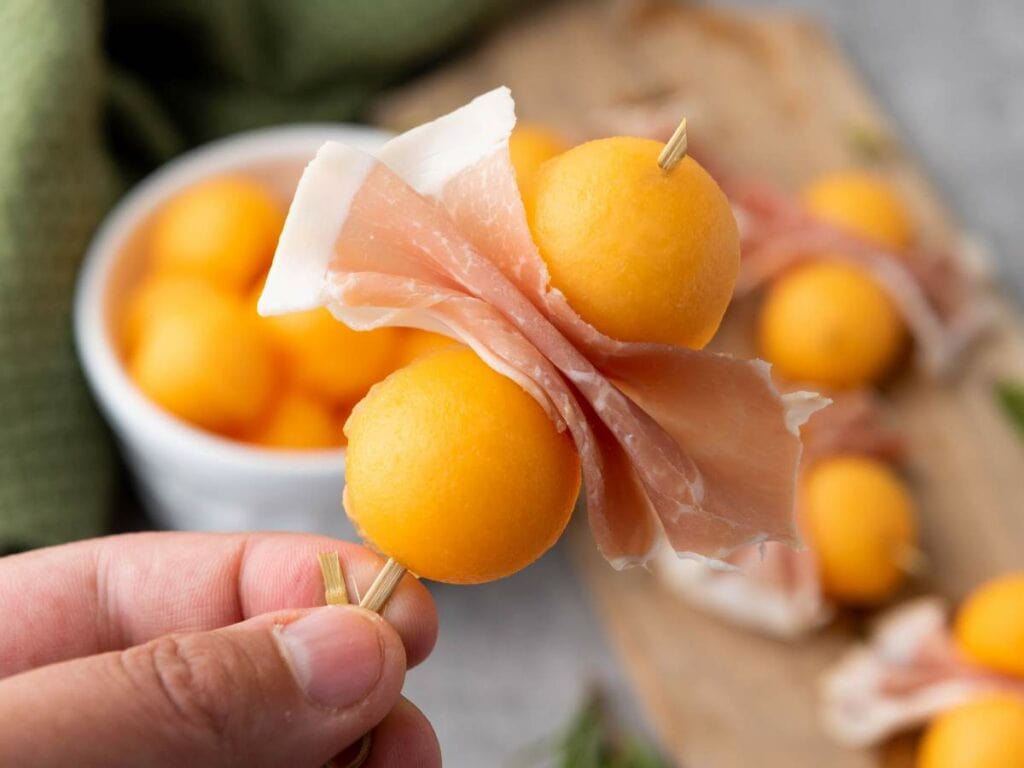 A hand holds a skewer with two prosciutto e melone and more skewers are visible in the background.