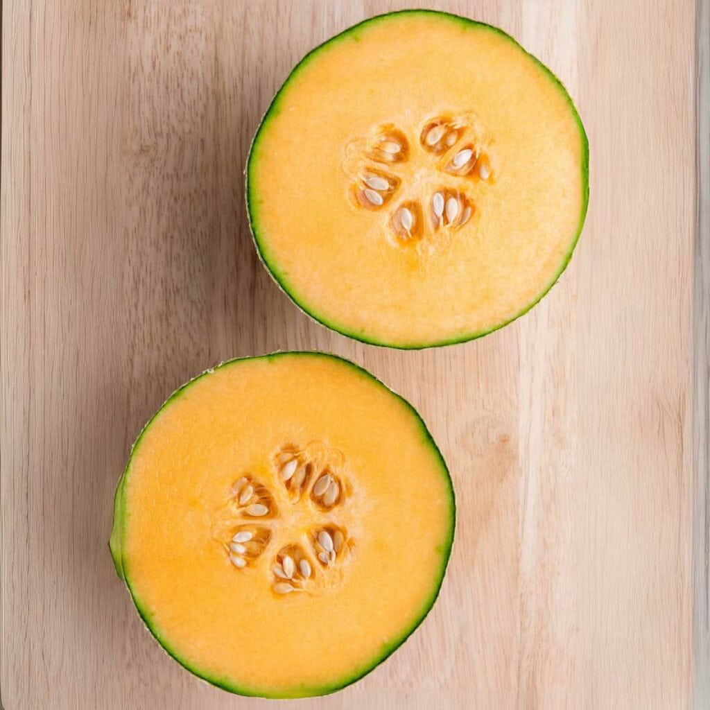 A cantaloupe cut in half, showing orange flesh and seeds, placed on a light wooden surface.