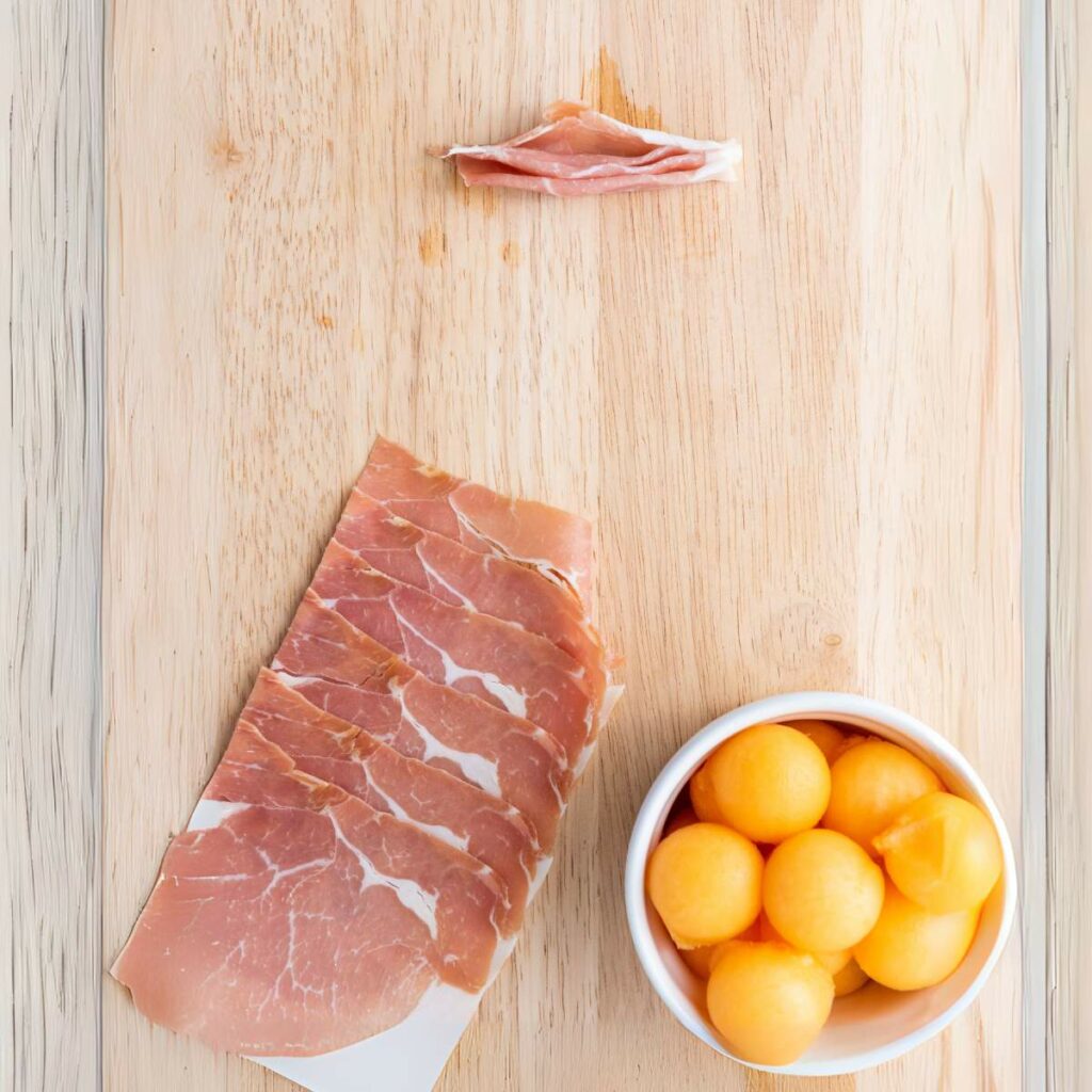 A wooden board with slices of prosciutto, a small bowl of melon balls, and a piece of prosciutto.