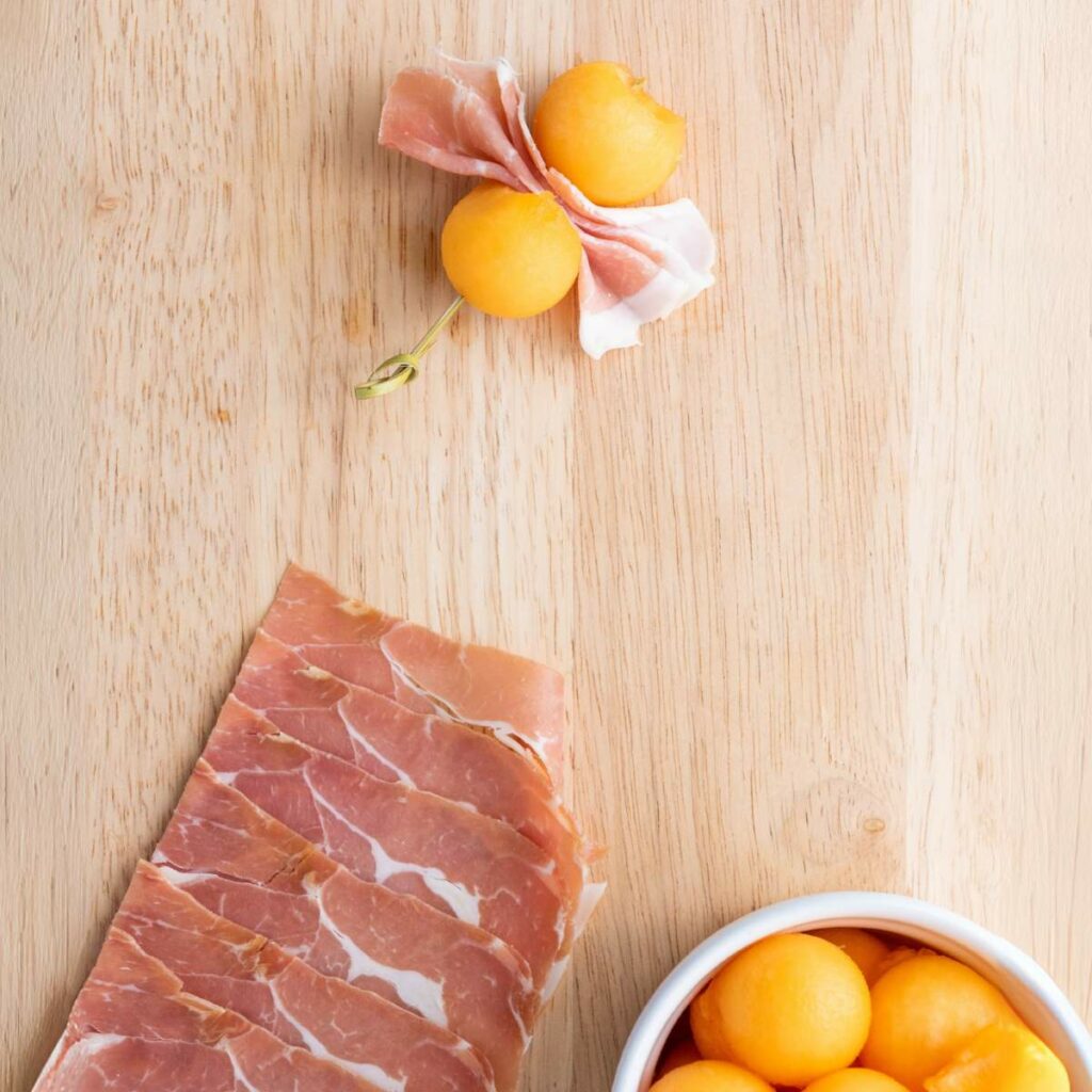 Slices of prosciutto, a bowl of melon balls, and a toothpick with melon balls wrapped in prosciutto.