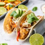 A chicken fajitas in a tortillas on a marble surface next to a pan of the same chicken fajitas.