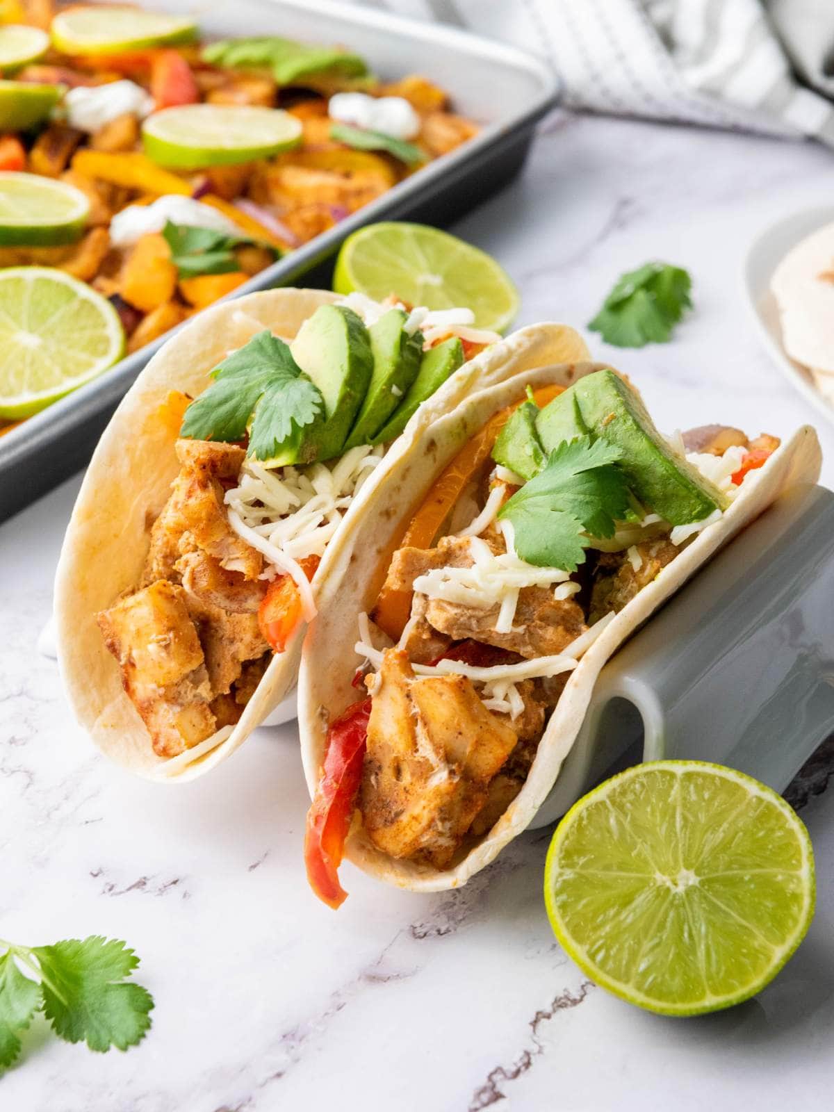 A chicken fajitas in a tortillas on a marble surface next to a pan of the same chicken fajitas.