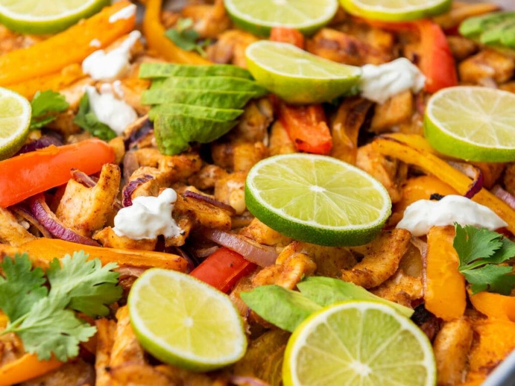 Close-up view of sheet pan chicken fajitas topped with sliced avocado, lime and fresh cilantro.
