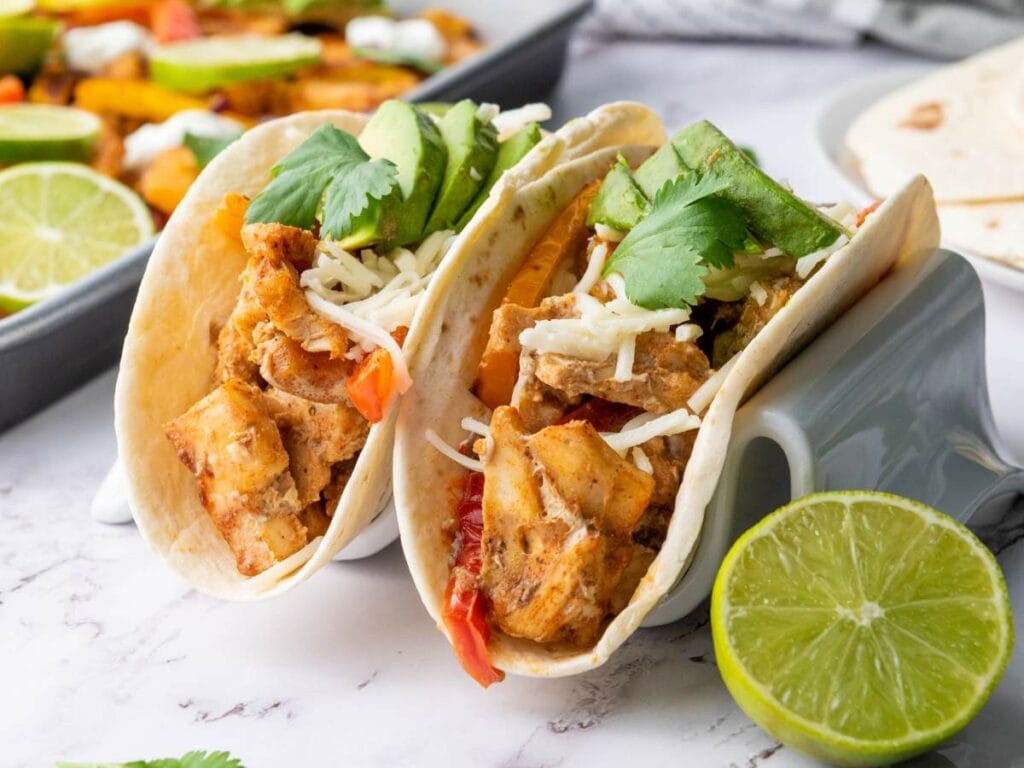 Two sheet pan chicken fajitas in a tortillas on a white taco holder and a half lemon on the side.