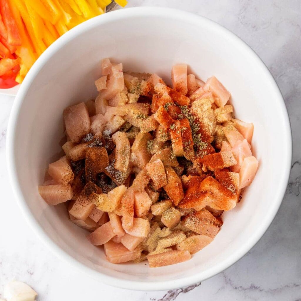 A white bowl containing sliced chicken breast with seasonings next to orange bell peppers.