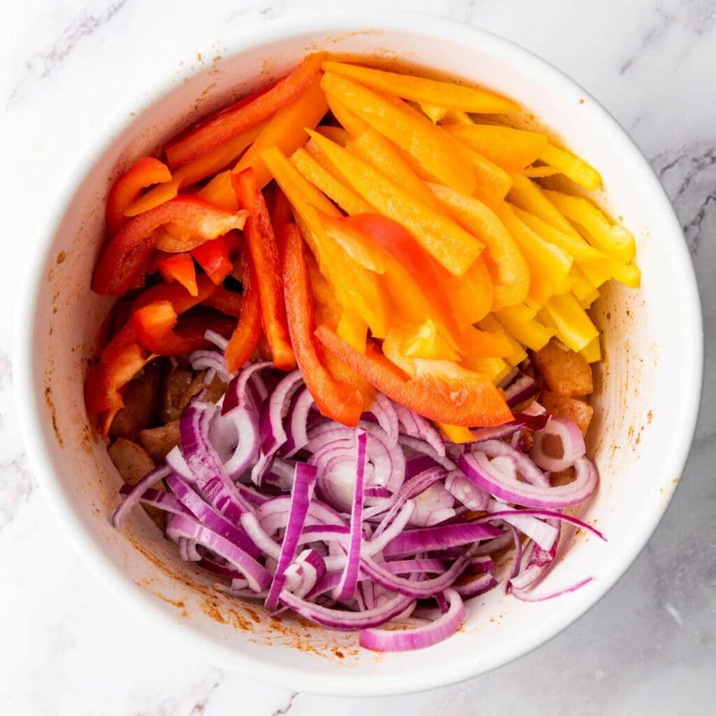 A bowl of seasoned chicken breast topped with sliced onions and bell peppers on a marble surface.