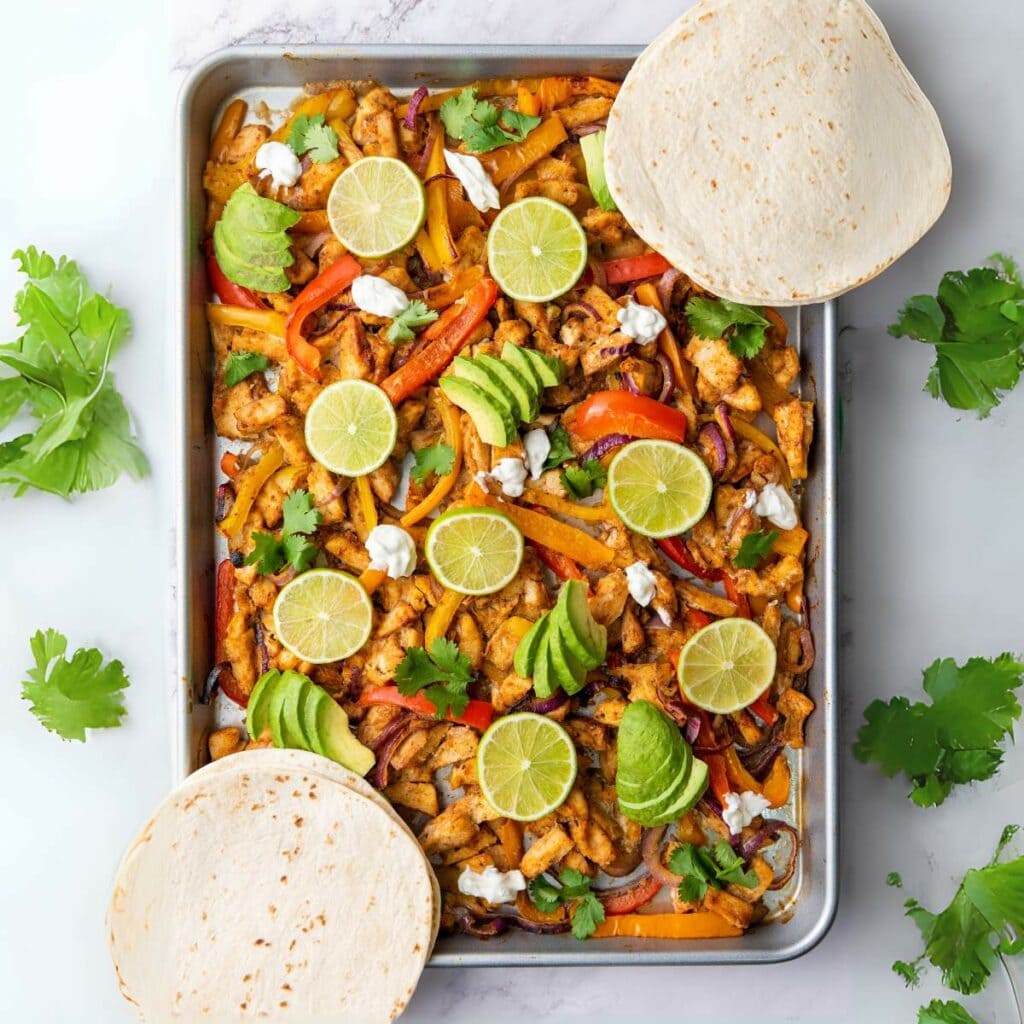 Cooked sheet pan chicken fajitas on a pan topped with sliced avocado and lime wedges with tortillas on the sides.