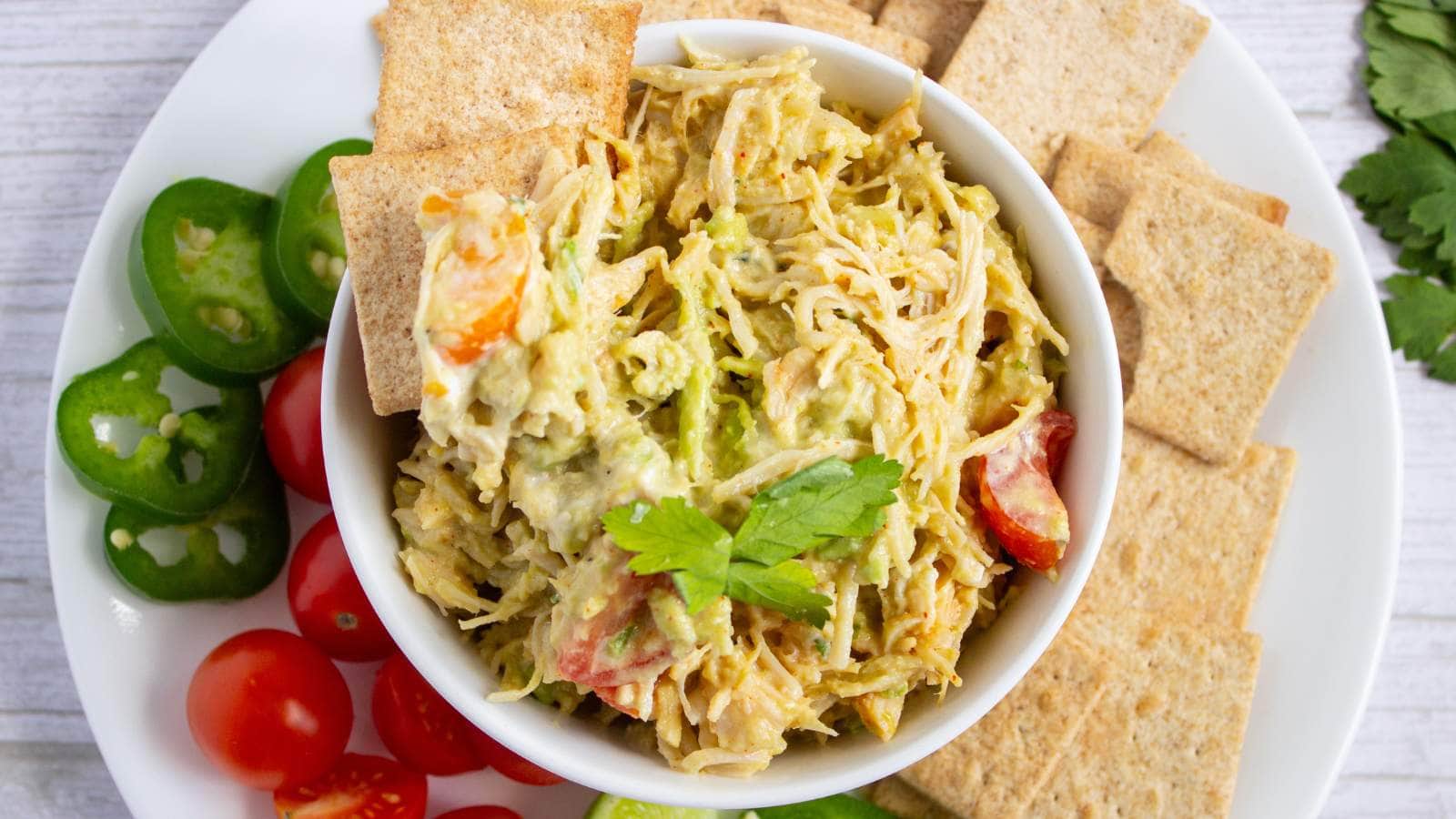 A white bowl of simple chicken salad recipe with tomatoes and herbs, surrounded by crackers, sliced jalapeños, and cherry tomatoes on a white plate.