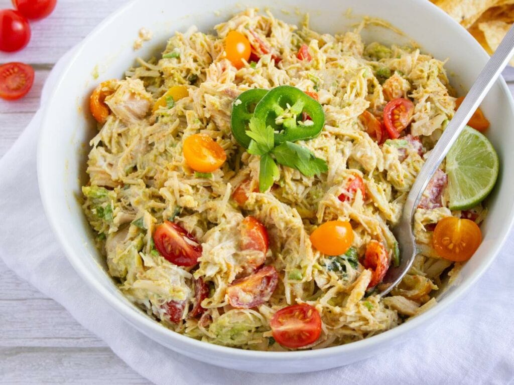 A bowl of simple chicken salad recipe garnished with a slice of lime and jalapeño, with a serving spoon on a white table napkin.