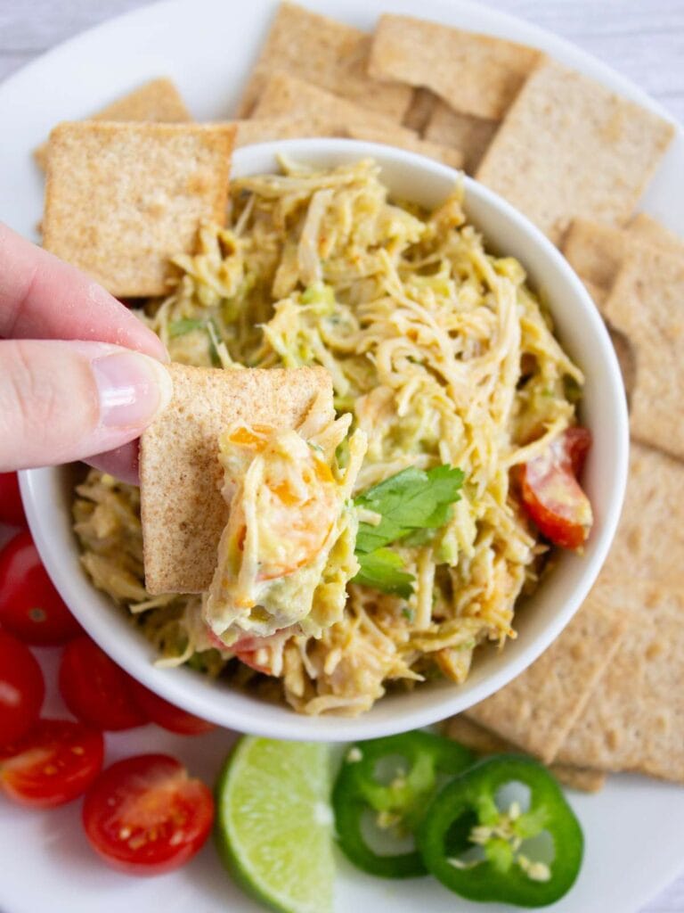 A hand dips a square cracker into a bowl of shredded chicken on a white plate.