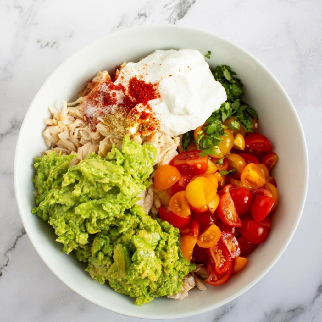 A white bowl containing shredded chicken, avocado, halved cherry tomatoes, chopped herbs, yogurt, and spices on a marble surface.