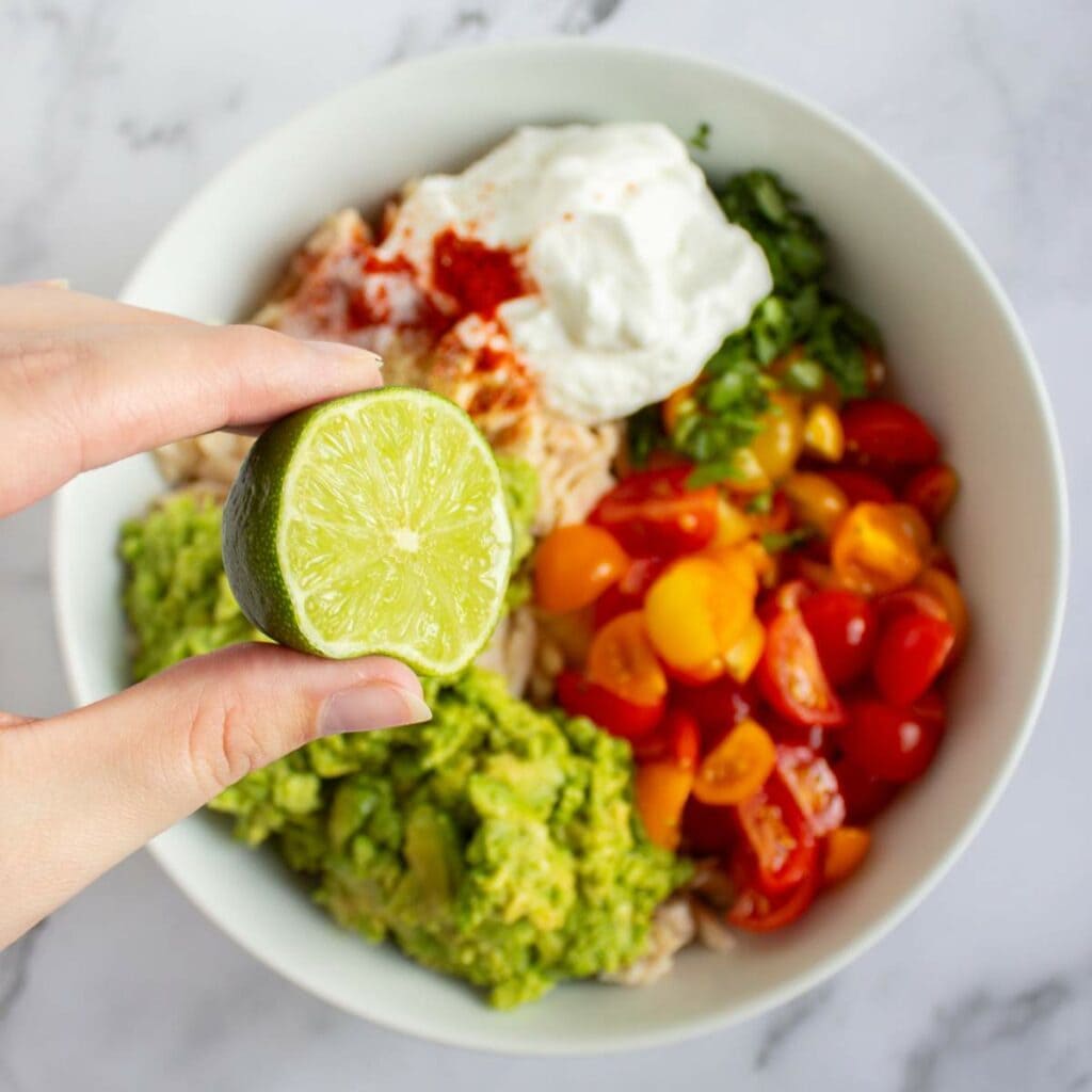 A hand holding half a lime over a bowl of mix ingredients of simple chicken salad on a marble surface.