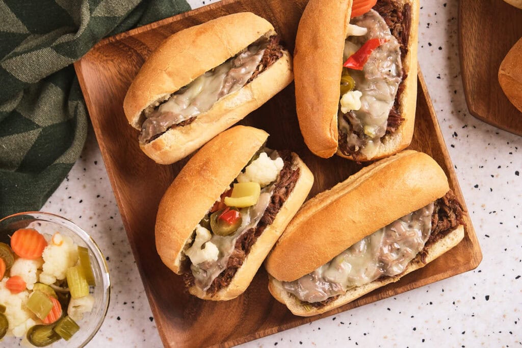 Four sandwiches filled with shredded italian beef, melted cheese, and topped with pickled vegetables are arranged on a wooden tray.
