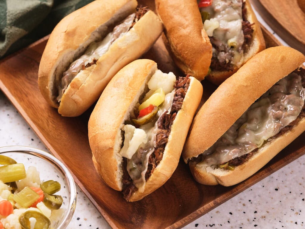 Slow cooker italian beef sandwiches with melted cheese and pickled vegetables on a wooden tray.
